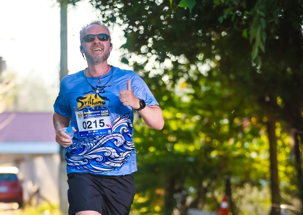 A runner in sunglasses and a blue wave-print race shirt smiles and gives a thumbs-up mid-stride on a tree-lined road, race number 0215 pinned to his chest and a water bottle in one hand